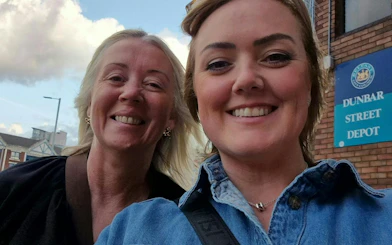 Two people smiling in front of Dunbar Street Depot sign, Belfast.
