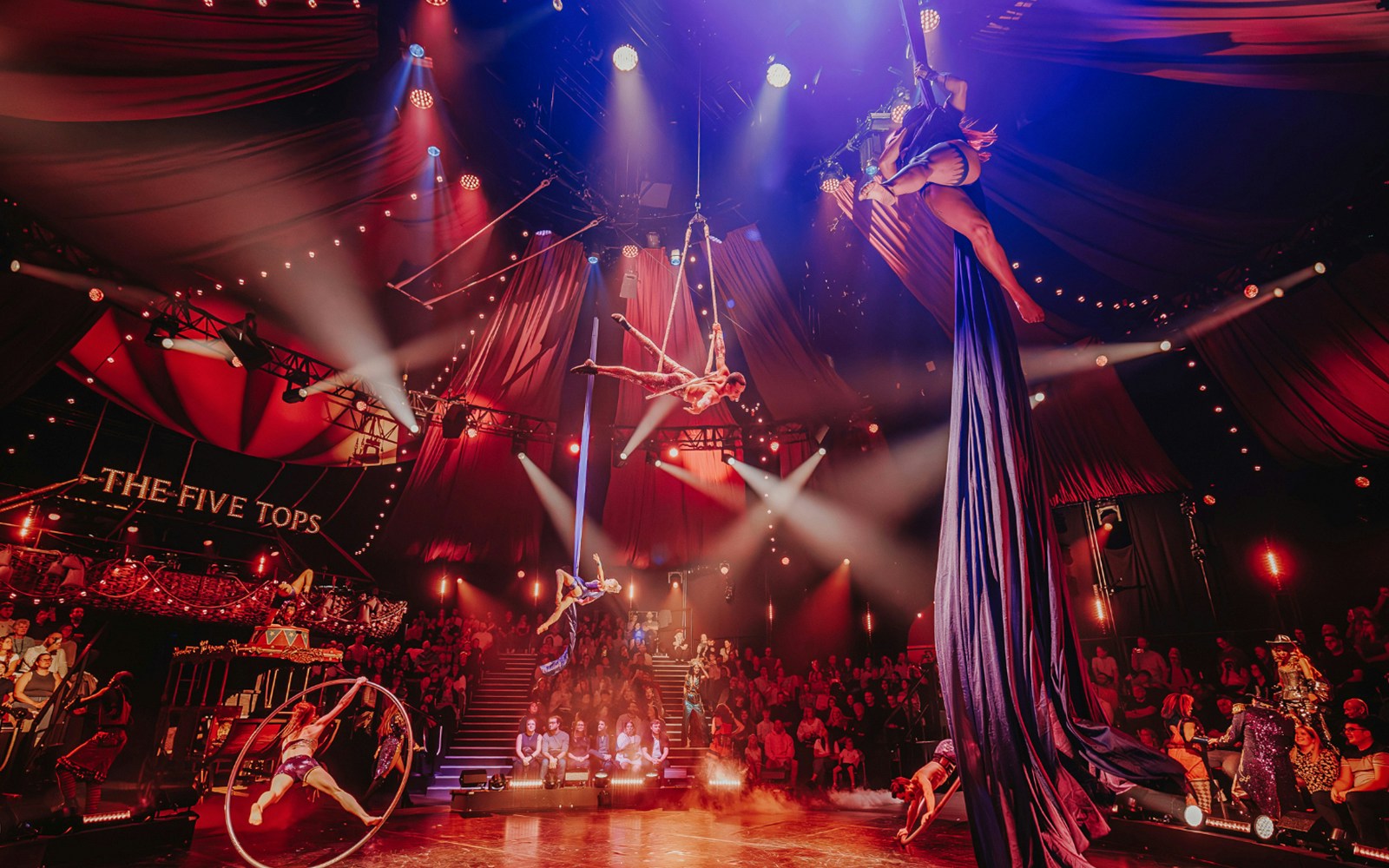 Aerial performers in a vibrant circus show with audience, titled "The Five Tops.