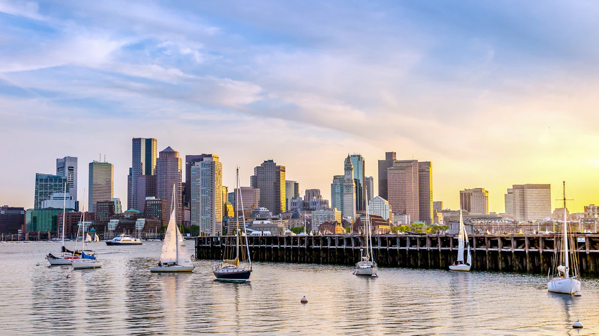 Sailboats on the water with the Boston skyline at sunset.