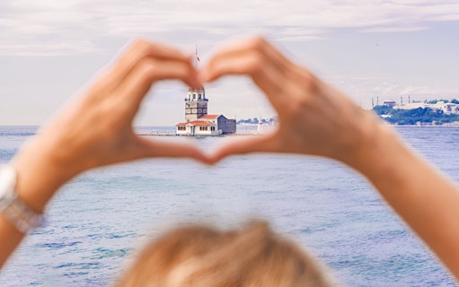 Hands forming a heart shape framing Maiden's Tower in Istanbul, viewed from the water.