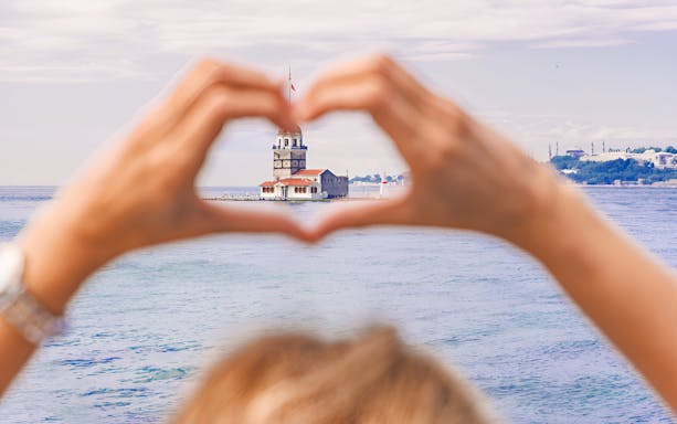Hands forming a heart shape framing Maiden's Tower in Istanbul, viewed from the water.