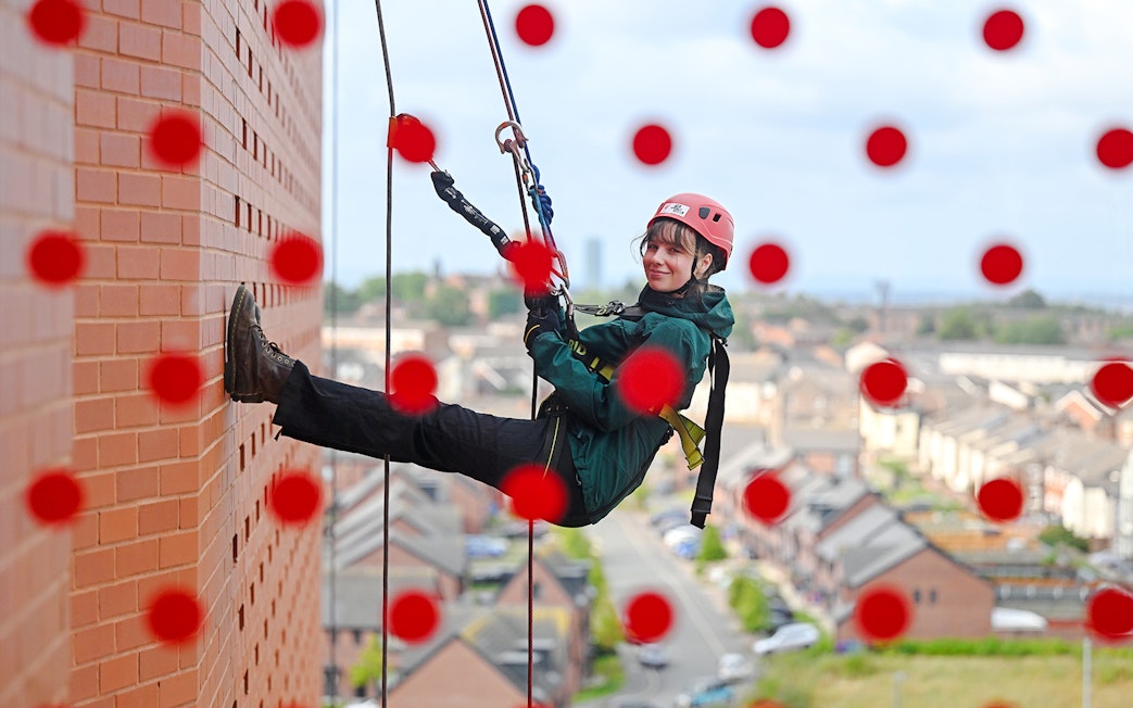 Guest abseiling down Anfield stadium wall with city view in background.
