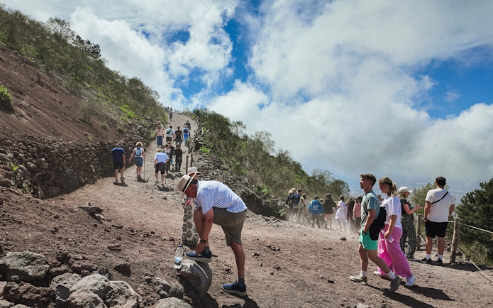Visitors hiking up Mount Vesuvius on a rocky path under a cloudy sky.