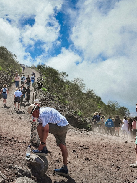 Visitors hiking up Mount Vesuvius on a rocky path under a cloudy sky.