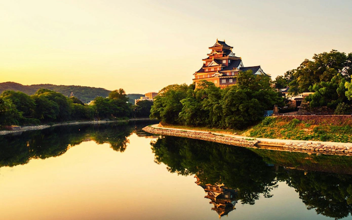Okayama Castle by a river at sunrise with a yellow sky in Okayama, Japan.