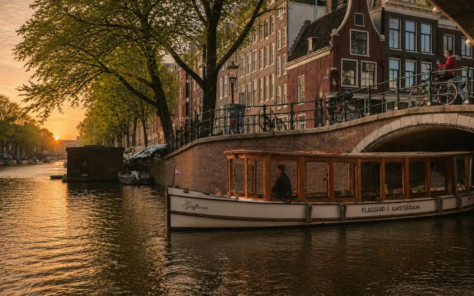 Luxury canal boat cruising under a bridge at sunset in Amsterdam.
