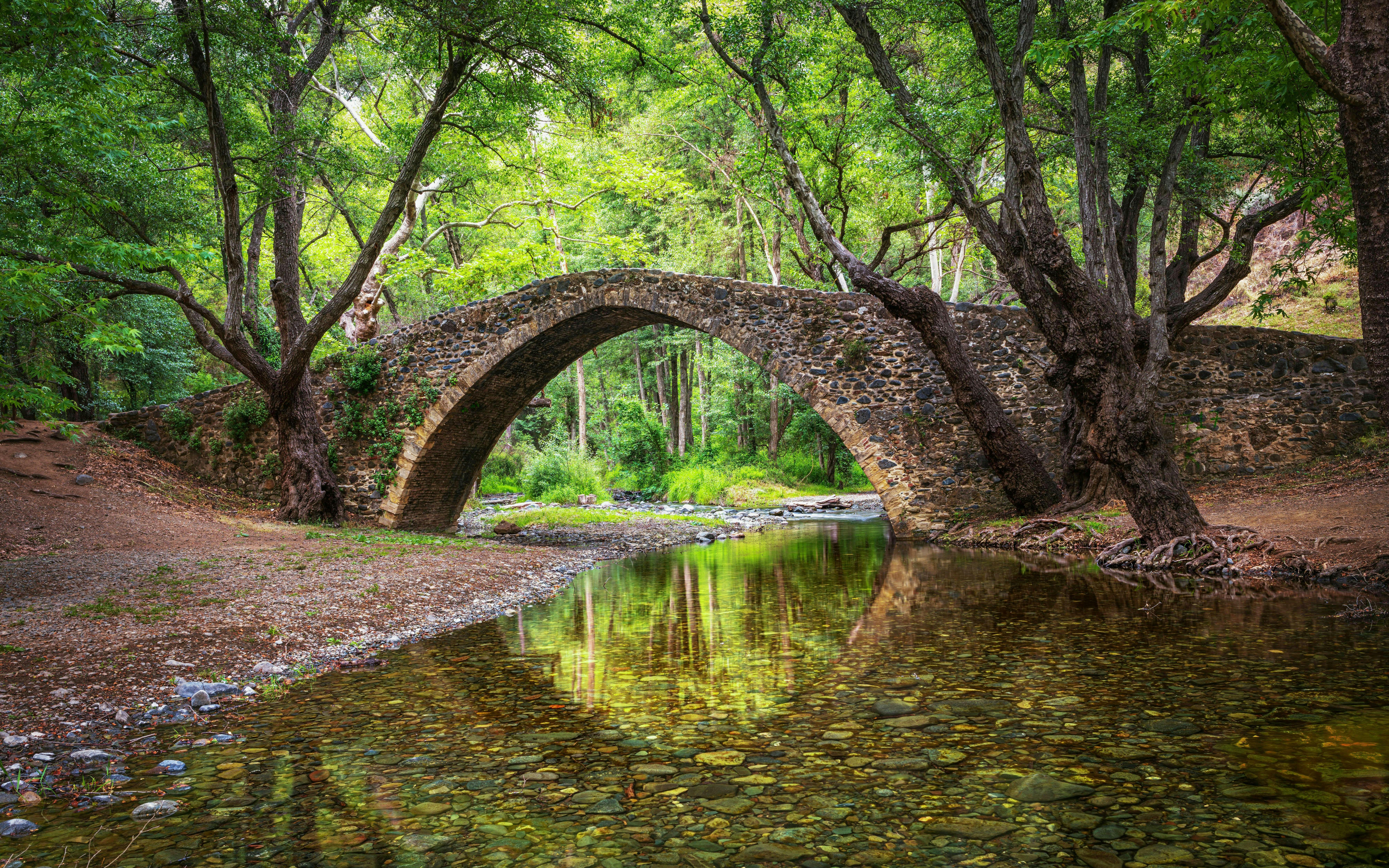 Kelefos Medieval Bridge in Cyprus reflected in a river, surrounded by lush trees.