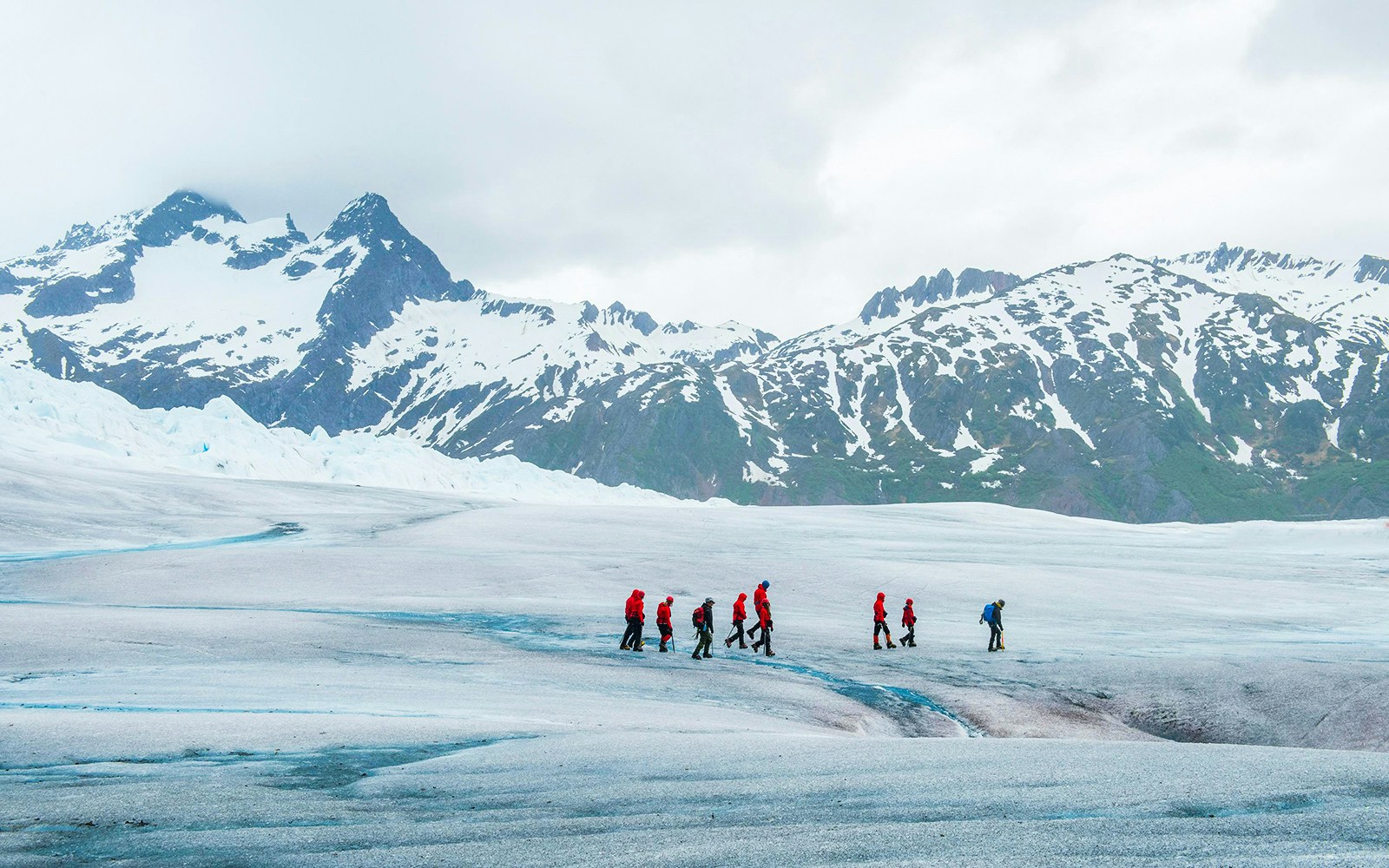 Hikers in red jackets walking on Mendenhall Glacier, Juneau, Alaska with snowy mountains in the background.