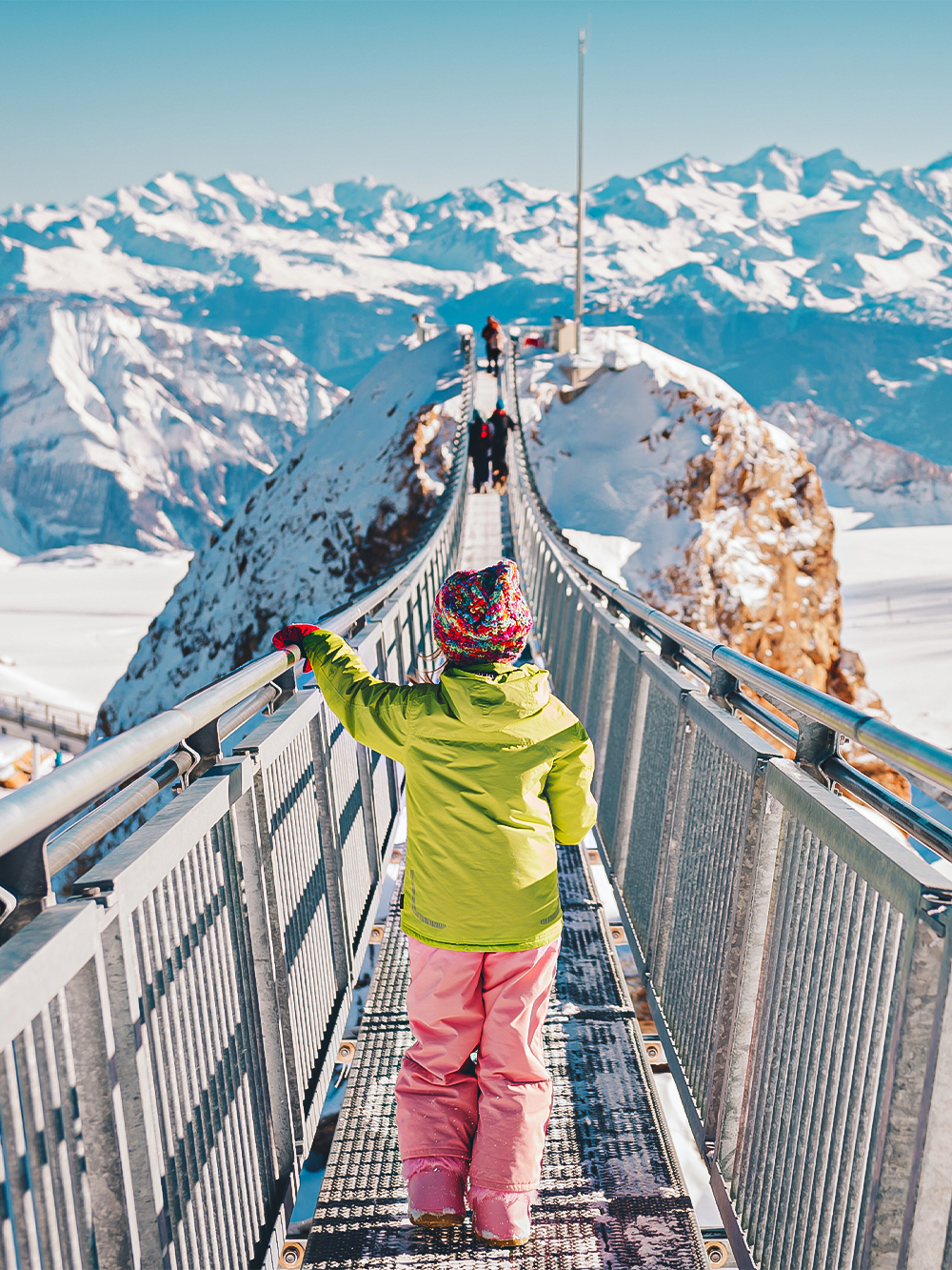 Child walking on Glacier 3000 suspension bridge with snowy Alps in background.