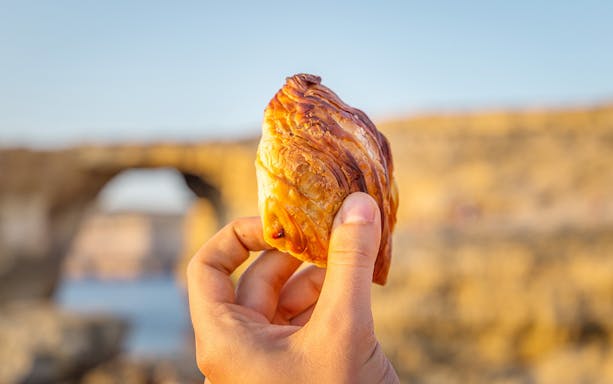 Hand holding a traditional Maltese pastizzi with a scenic view in Mdina.