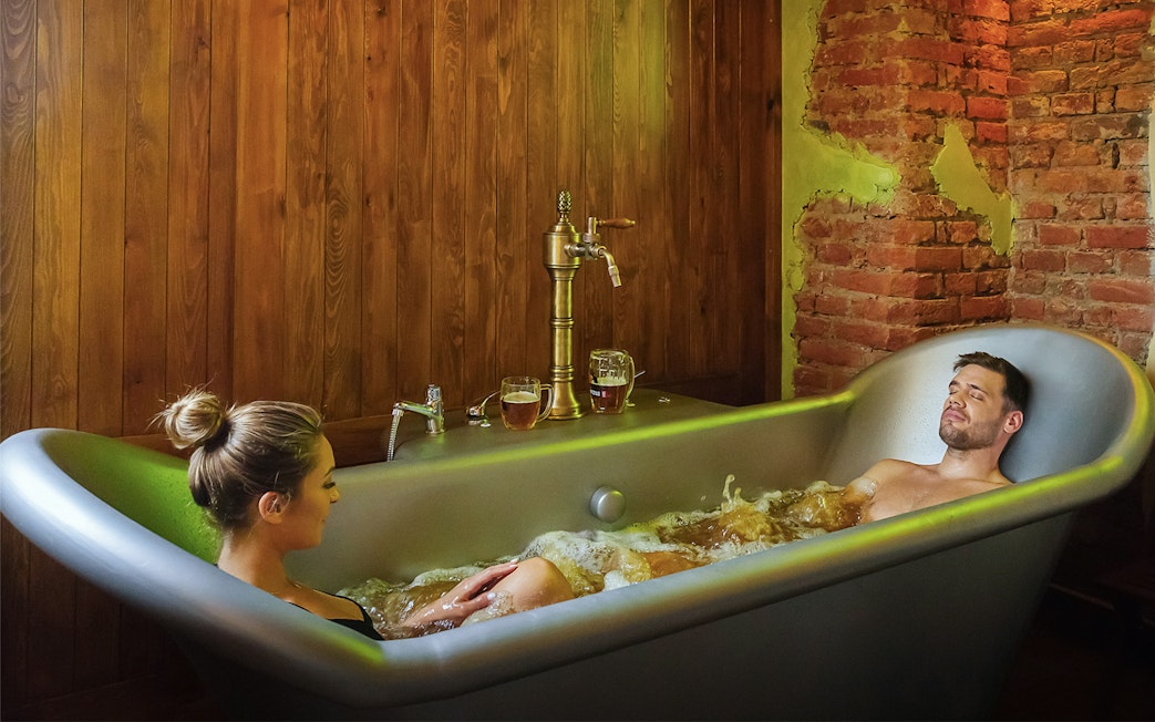 Couple relaxing in a beer spa bath with wooden and brick decor.