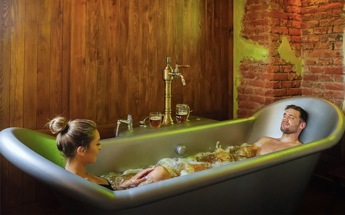 Couple relaxing in a beer spa bath with wooden and brick decor.