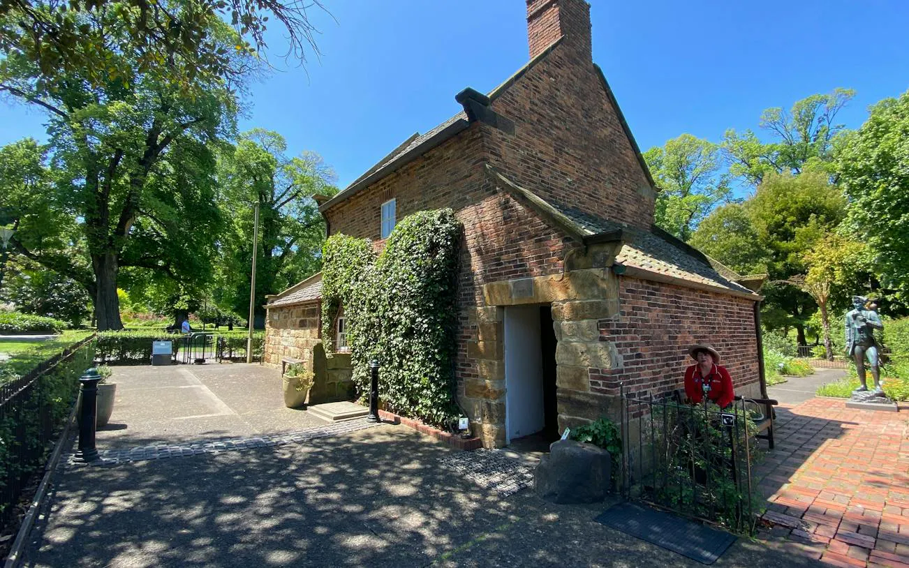 Historic cottage in Fitzroy Gardens, Melbourne, surrounded by lush greenery and a statue nearby.