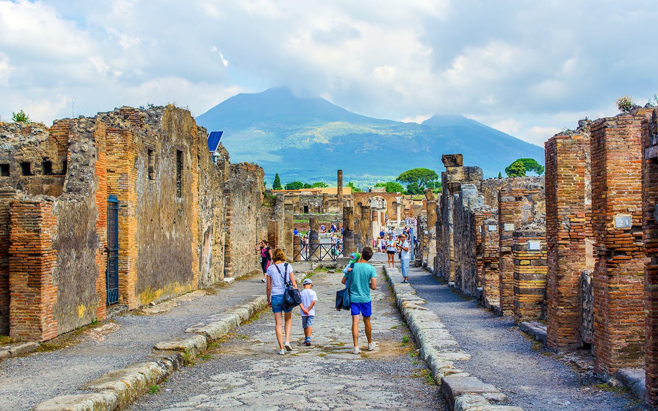 Tourists walking through ancient ruins of Pompeii with Mount Vesuvius in the background.