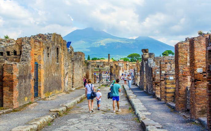 Tourists walking through ancient ruins of Pompeii with Mount Vesuvius in the background.