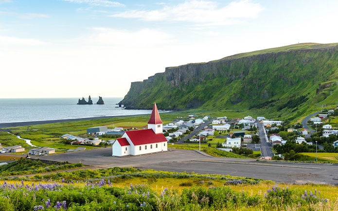 Vik church with red roof, coastal cliffs, and Reynisdrangar sea stacks in Vik, Iceland.