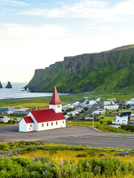 Vik church with red roof, coastal cliffs, and Reynisdrangar sea stacks in Vik, Iceland.