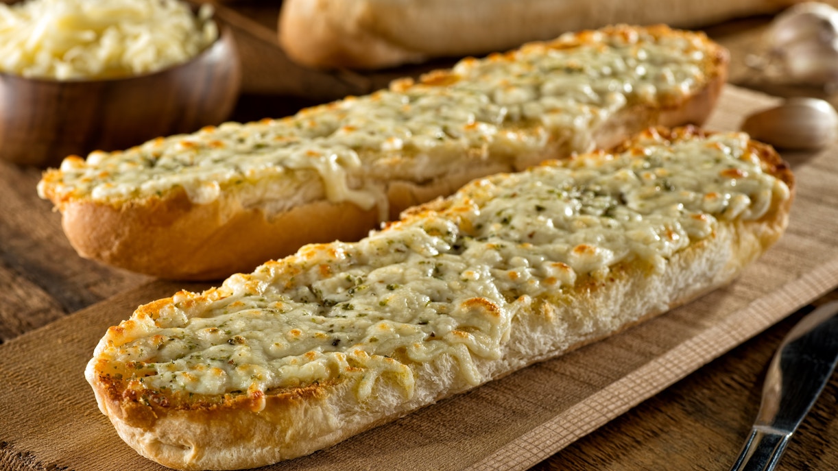 Cheesy garlic bread on a wooden board with garlic cloves nearby.