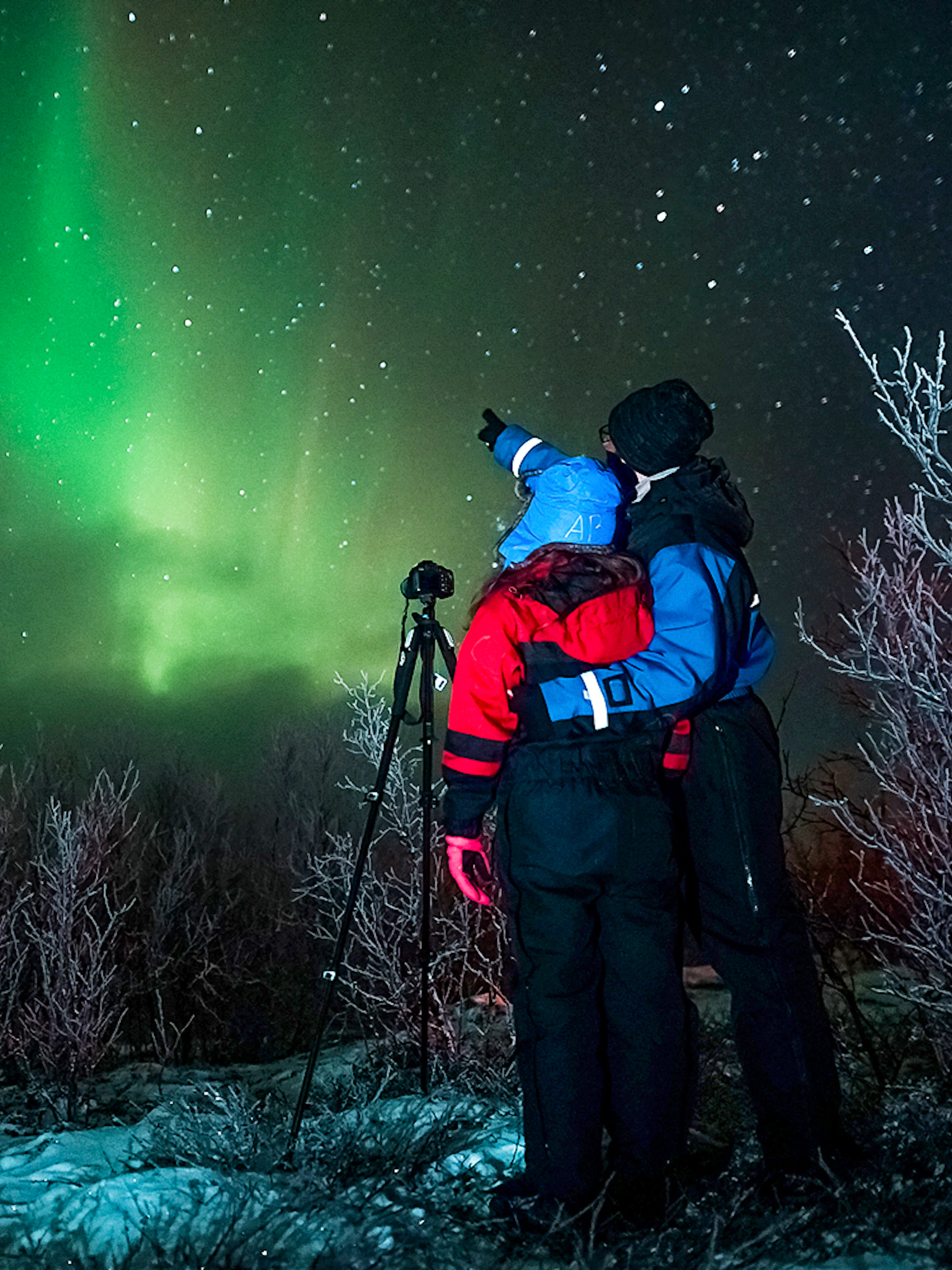 Tour guide with camera gear pointing at northern lights in snowy landscape.