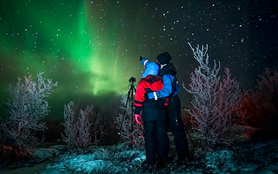 Tour guide with camera gear pointing at northern lights in snowy landscape.