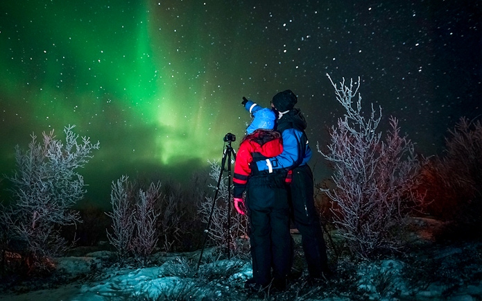Tour guide with camera gear pointing at northern lights in snowy landscape.
