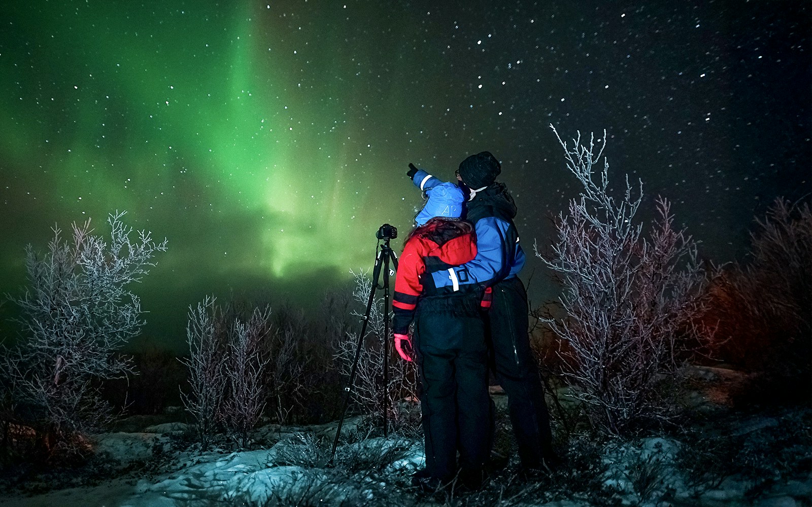 Tour guide with camera gear pointing at northern lights in snowy landscape.