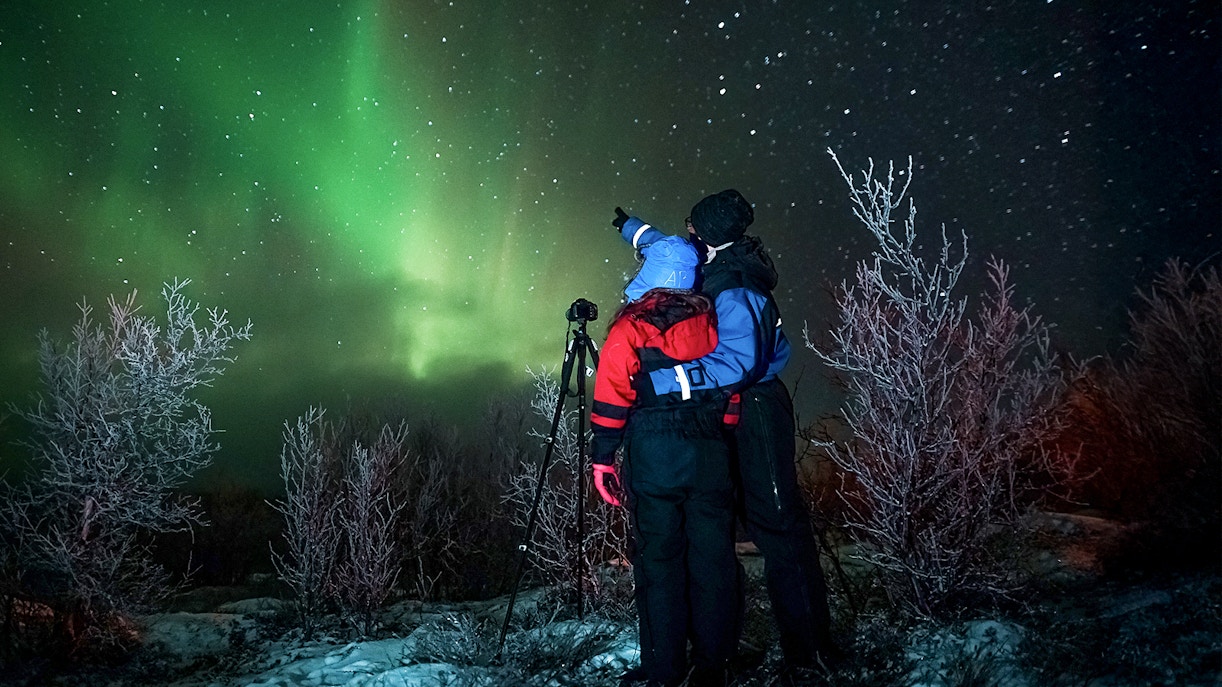 Tour guide with camera gear pointing at northern lights in snowy landscape.