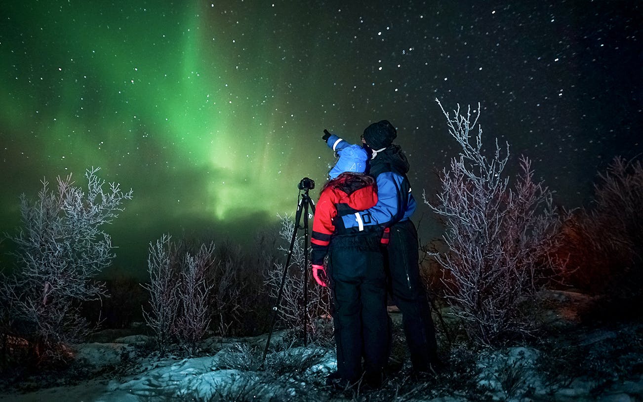 Tour guide with camera gear pointing at northern lights in snowy landscape.