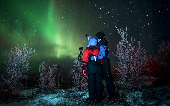 Tour guide with camera gear pointing at northern lights in snowy landscape.