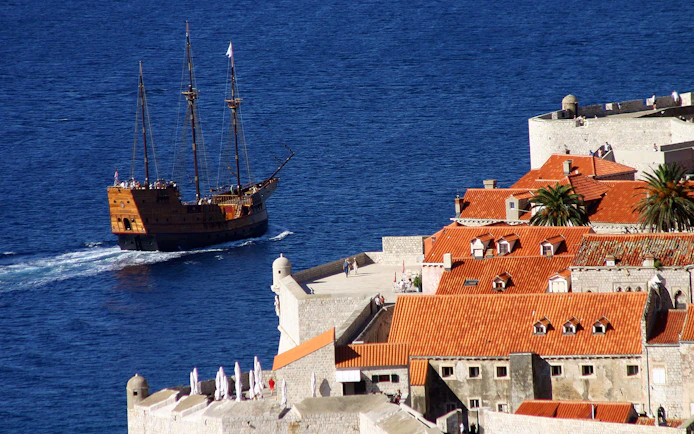Cruise ship sailing near Dubrovnik's historic city walls and red-roofed buildings.