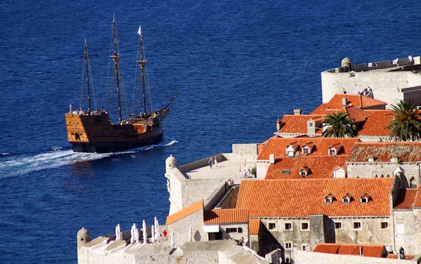 Cruise ship sailing near Dubrovnik's historic city walls and red-roofed buildings.