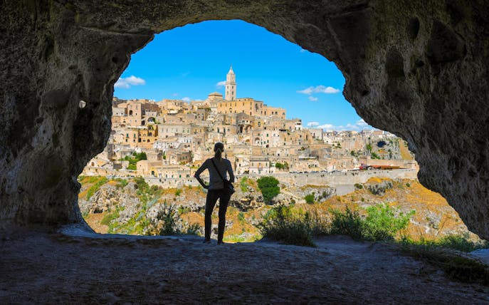 Woman viewing Matera skyline from cave across canyon, Italy.
