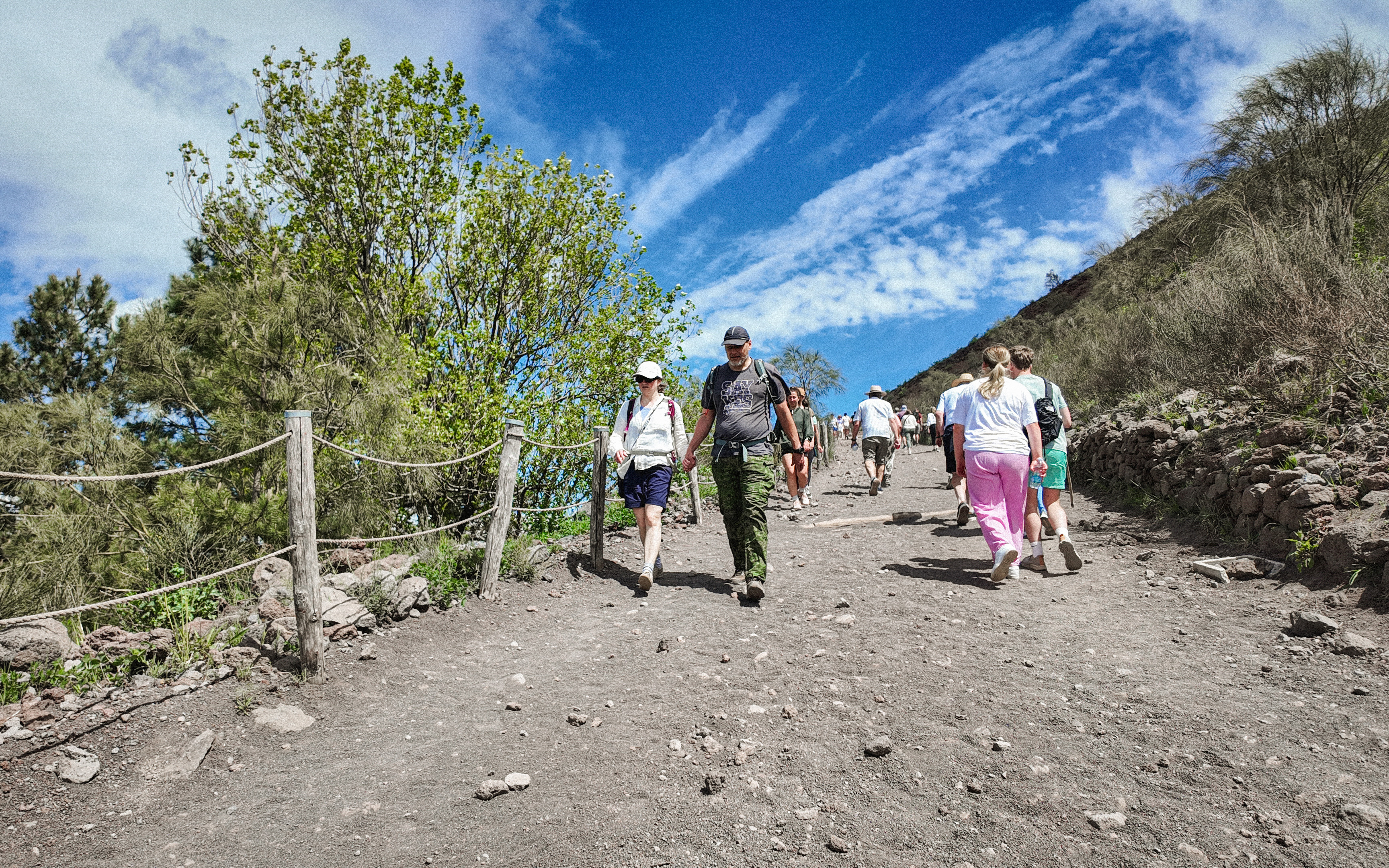 Visitors hiking up Mount Vesuvius trail surrounded by greenery and blue sky.