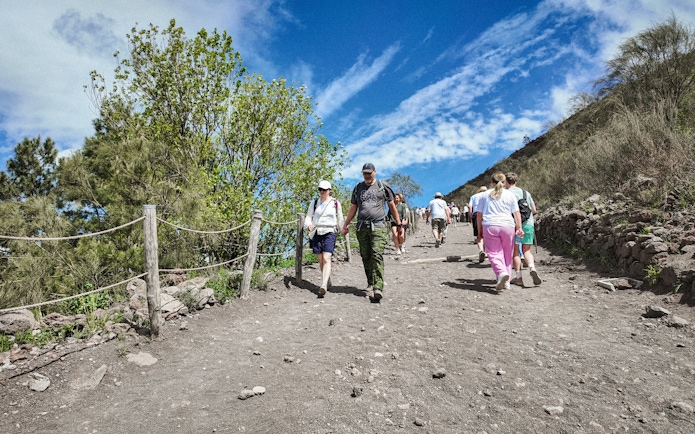 Visitors hiking up Mount Vesuvius trail surrounded by greenery and blue sky.