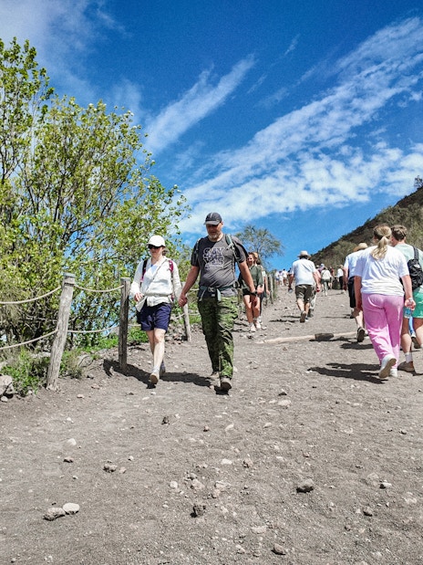 Visitors hiking up Mount Vesuvius trail surrounded by greenery and blue sky.