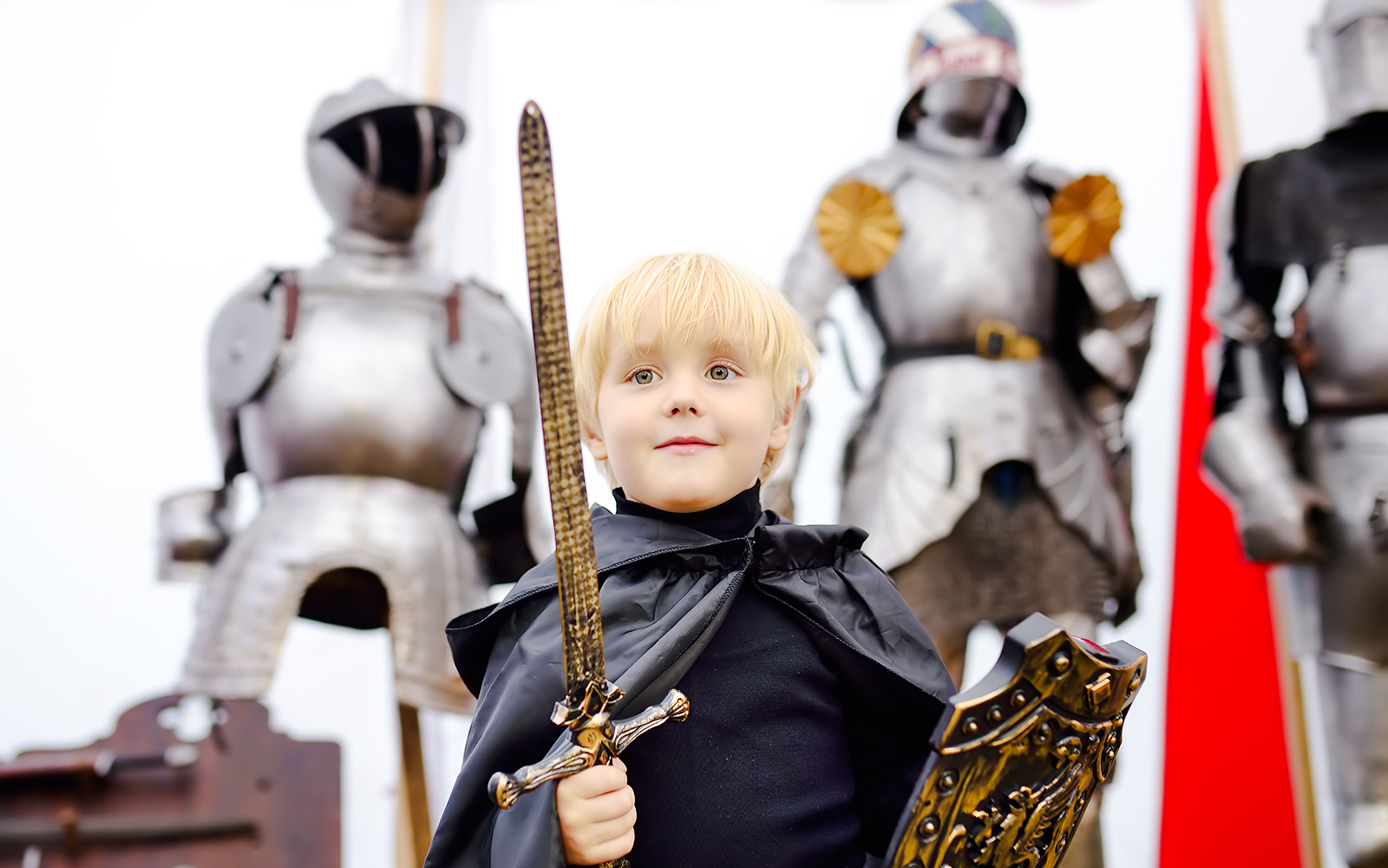 Child in medieval knight costume holding sword and shield, with armor display in background.