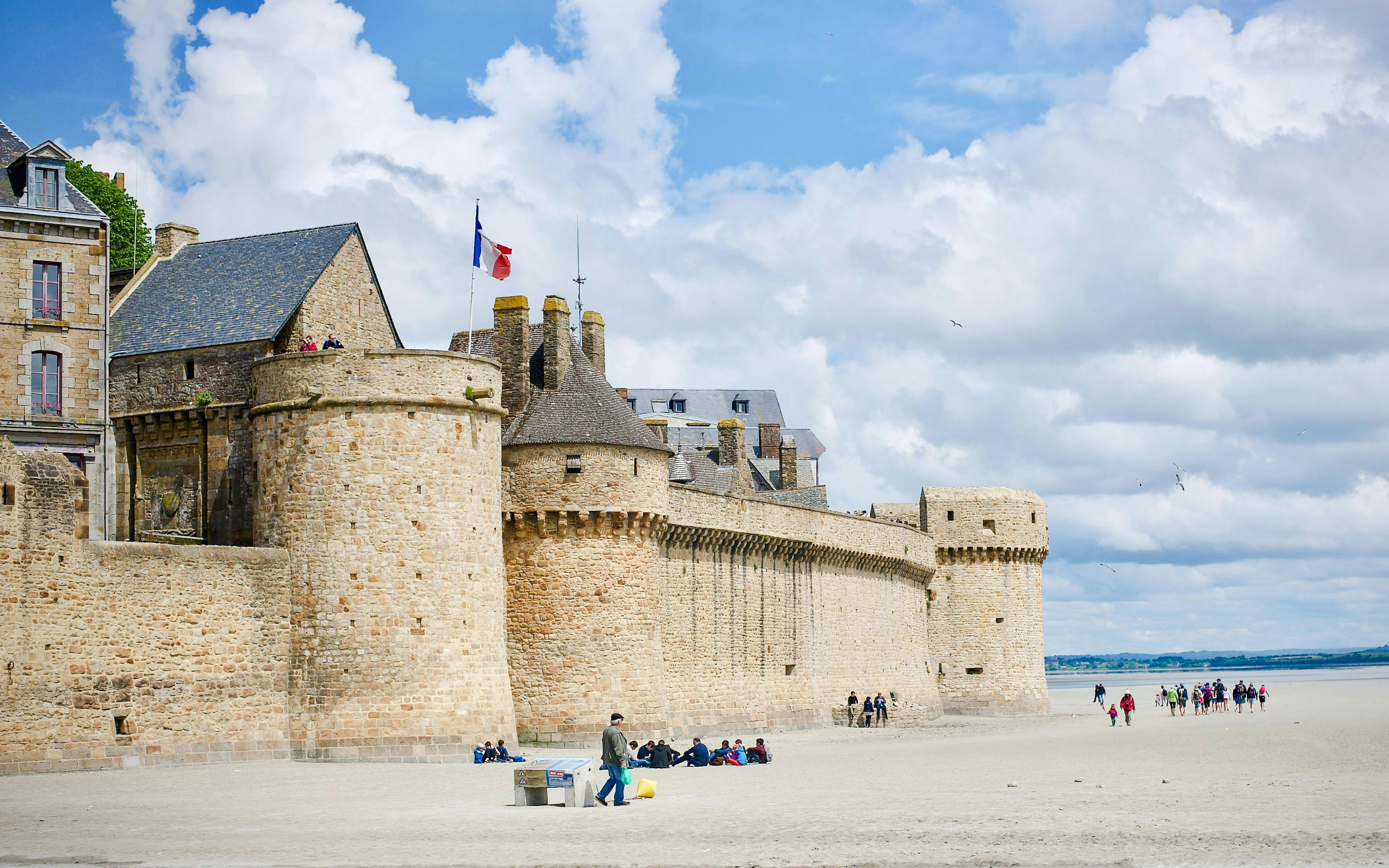 Ramparts of Mont-Saint-Michel Abbey with visitors walking on the sandy beach.