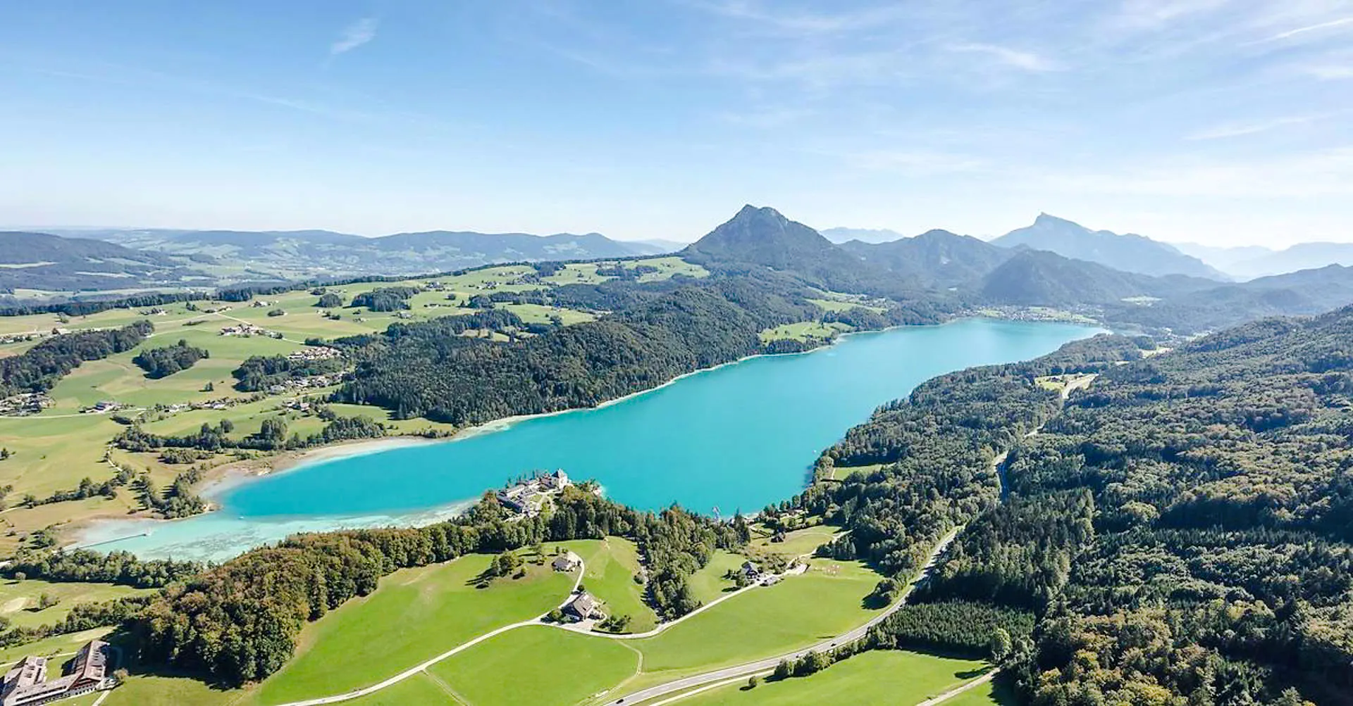 Aerial view of turquoise Alpine lake surrounded by lush greenery and mountains near Salzburg.