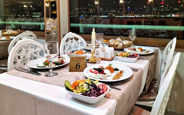 Elegant dinner setup with diverse dishes at a waterfront restaurant, city lights visible through the window.