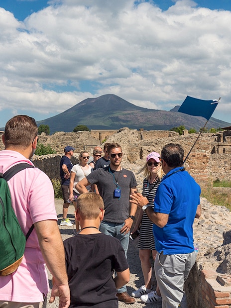 Guide explaining Pompeii ruins to tourists with Mount Vesuvius in the background.