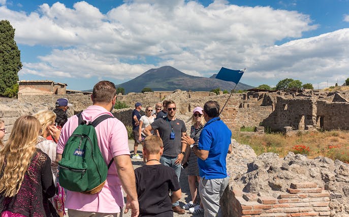 Guide explaining Pompeii ruins to tourists with Mount Vesuvius in the background.
