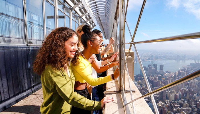 Tourists viewing New York City skyline from Empire State Building observation deck.