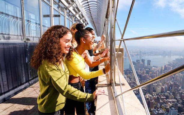 Tourists viewing New York City skyline from Empire State Building observation deck.