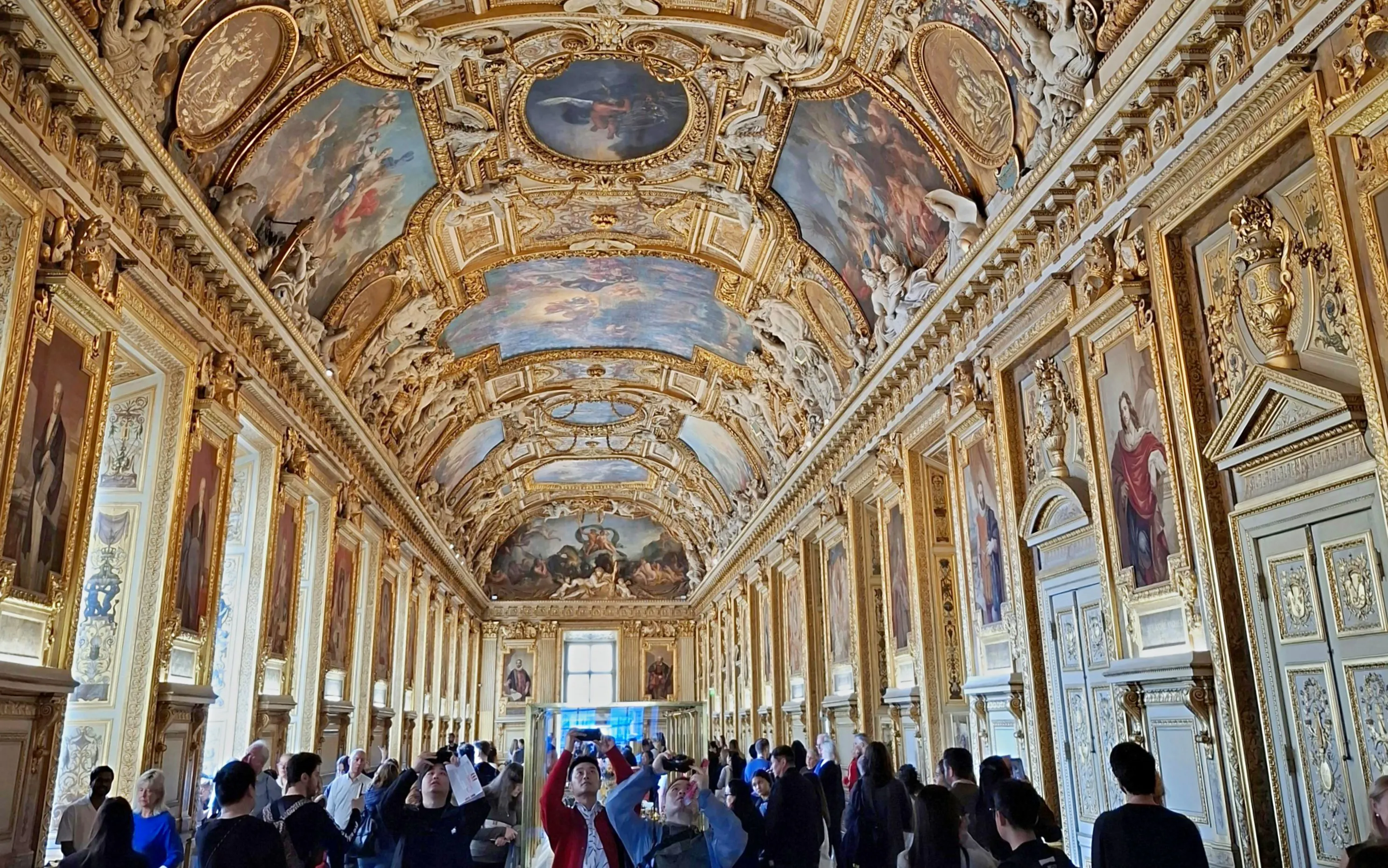 Visitors exploring the ornate Apollo Gallery in the Louvre, Paris, with detailed ceiling artwork.