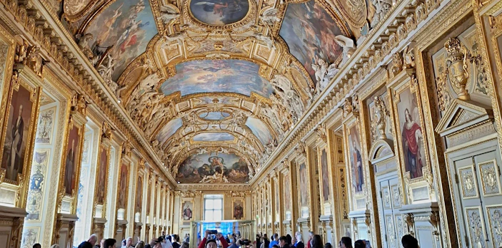 Visitors exploring the ornate Apollo Gallery in the Louvre, Paris, with detailed ceiling artwork.