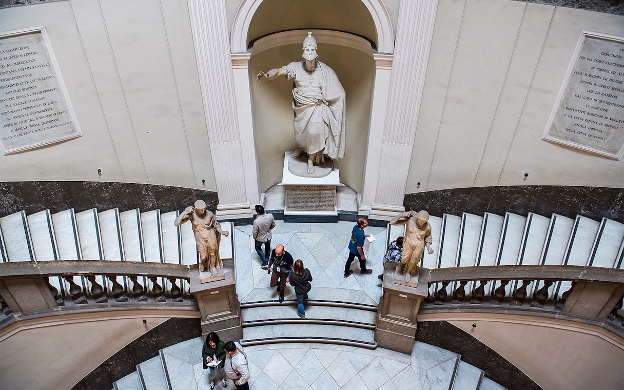 Visitors exploring statues and grand staircase at Archaeological Museum of Naples.