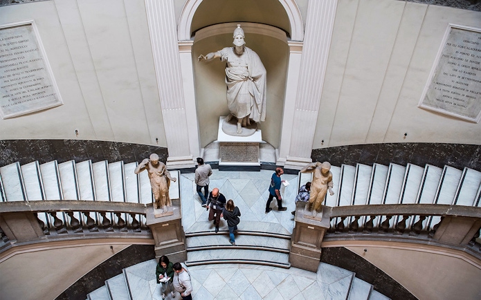 Visitors exploring statues and grand staircase at Archaeological Museum of Naples.