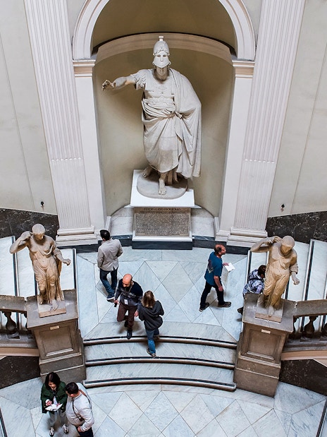 Visitors exploring statues and grand staircase at Archaeological Museum of Naples.