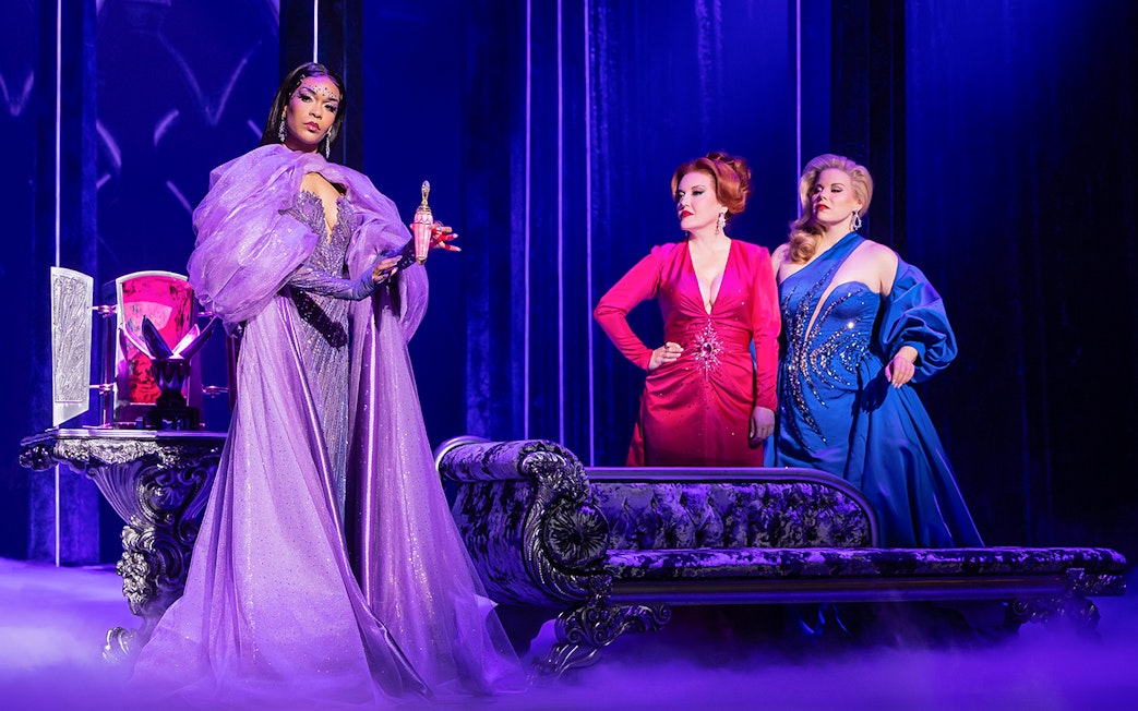 Three women in elegant gowns on stage, one holding a glowing potion, in "Death Becomes Her" performance.