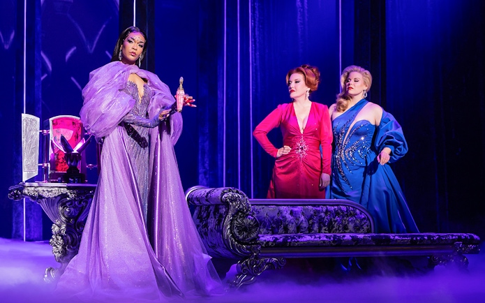 Three women in elegant gowns on stage, one holding a glowing potion, in "Death Becomes Her" performance.